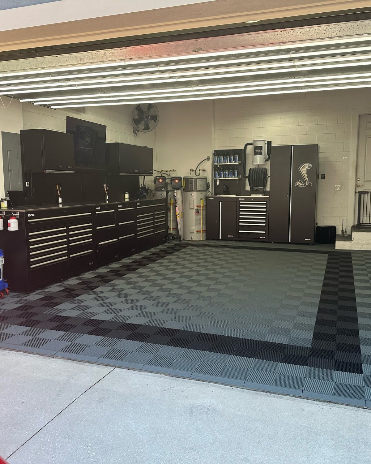 Organized garage with black tool cabinets, workbenches, and a checkered grey and black floor mat.