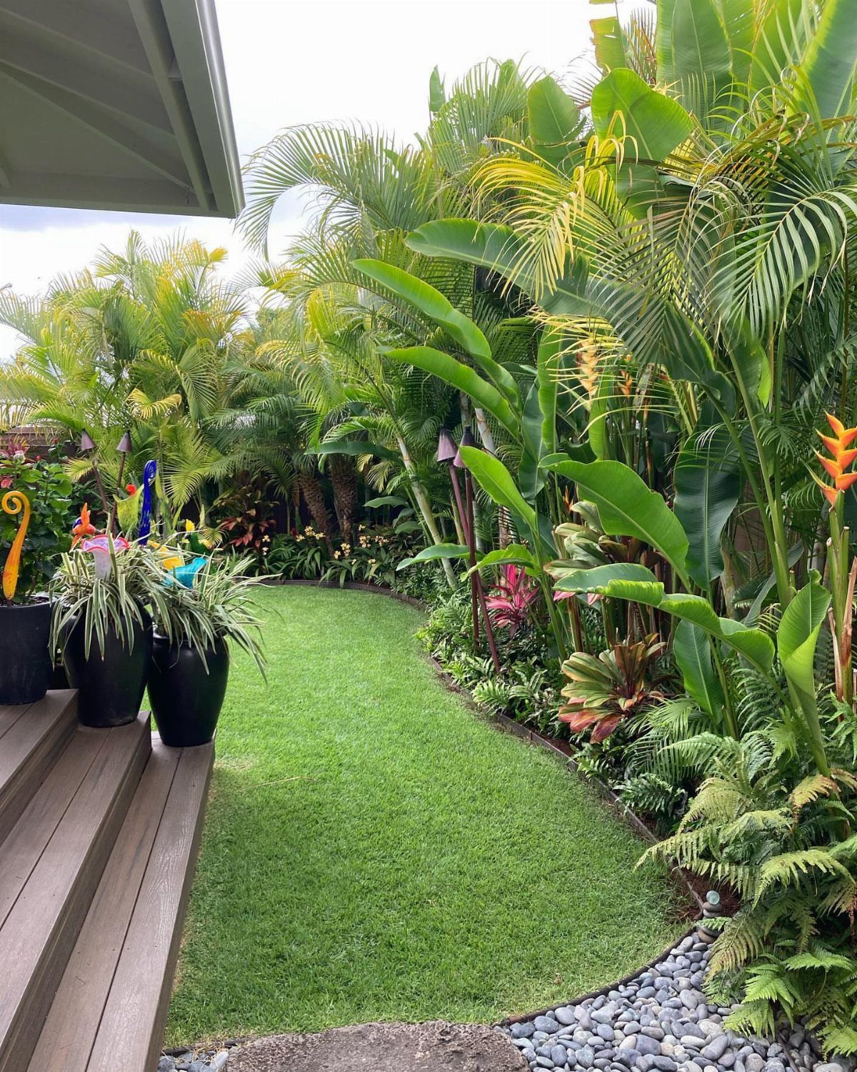 Lush tropical backyard garden with a green lawn path, palm trees, and decorative potted plants.
