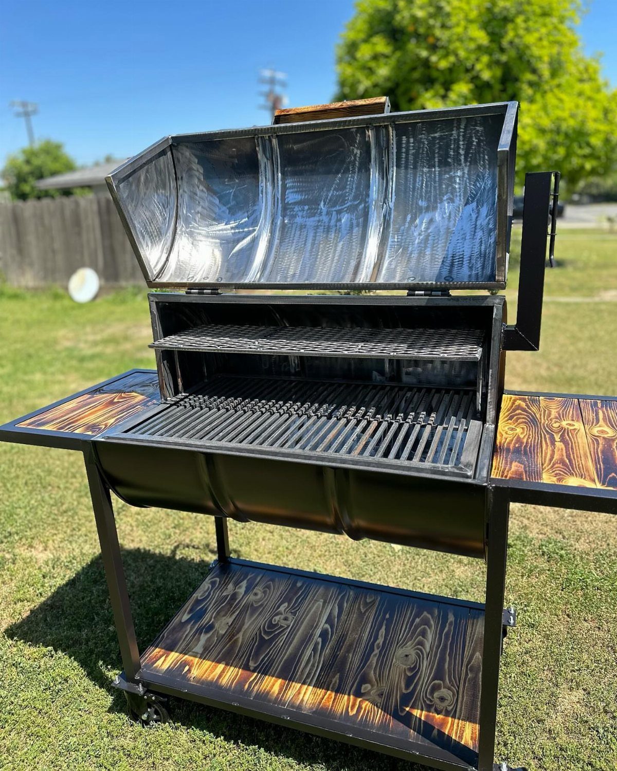 A custom black metal drum grill with wood-grain patterned side tables sitting on green grass.
