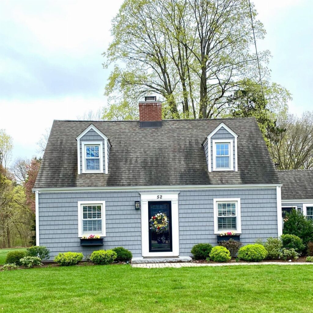 A grey cedar shake Cape Cod house with dormer windows and a green lawn.