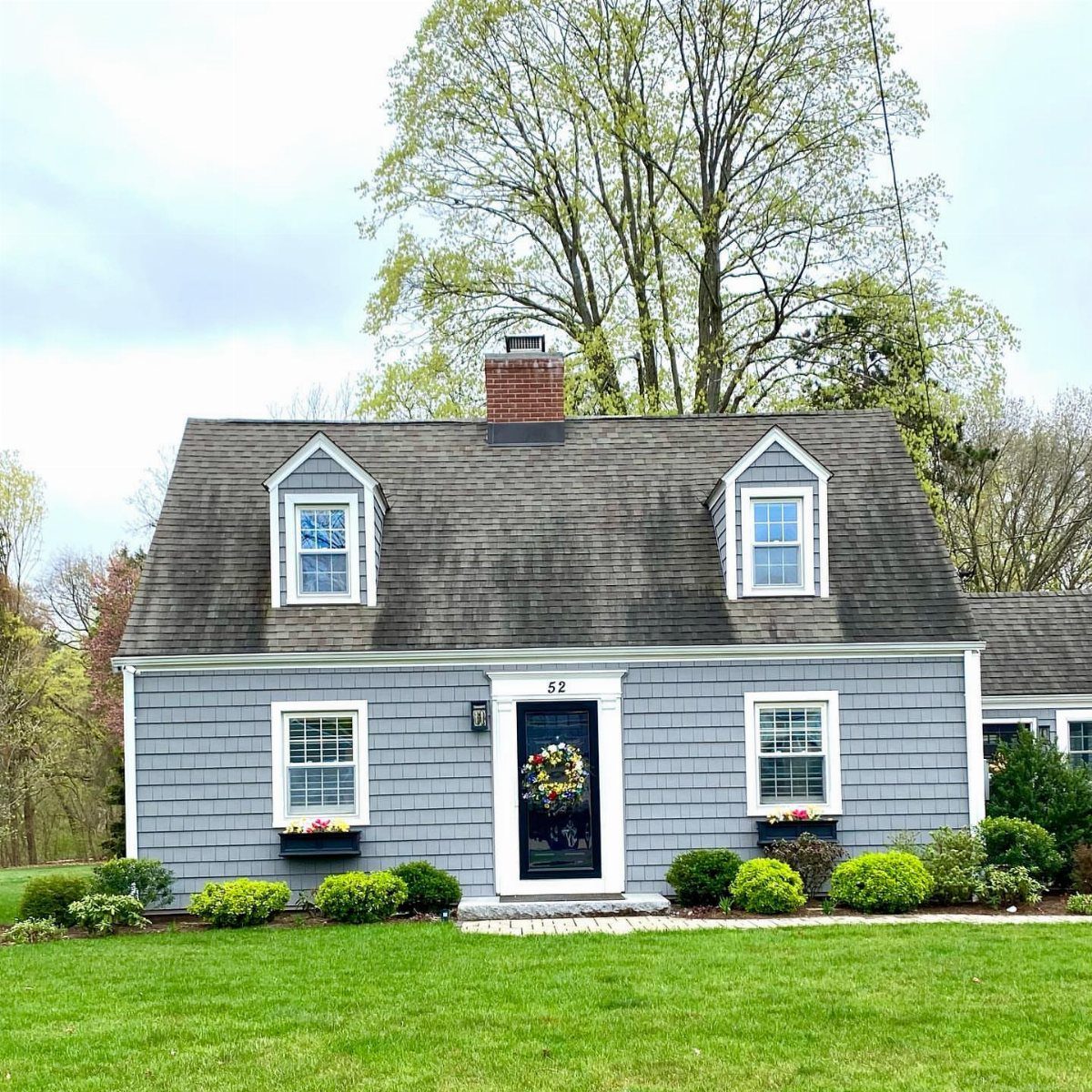 A grey cedar shake Cape Cod house with dormer windows and a green lawn.