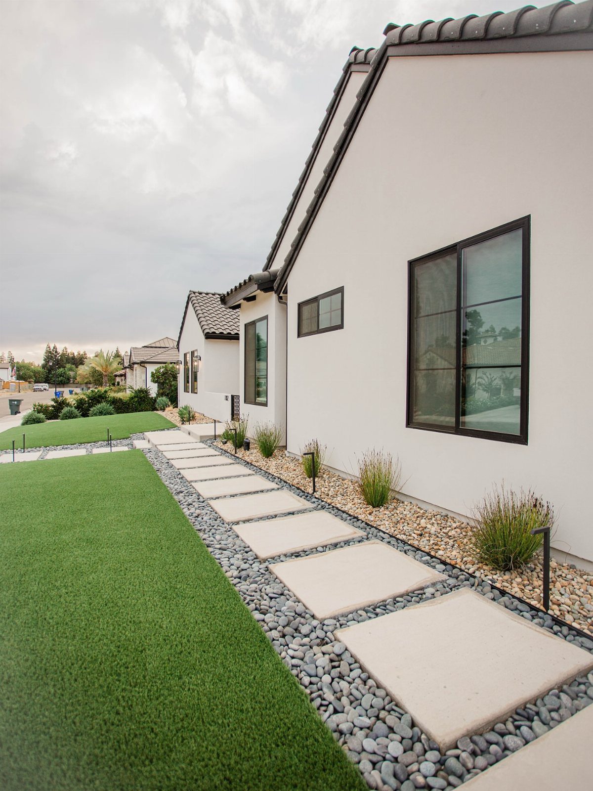 Concrete pavers set in grey stones create a walkway alongside a green lawn and white house.