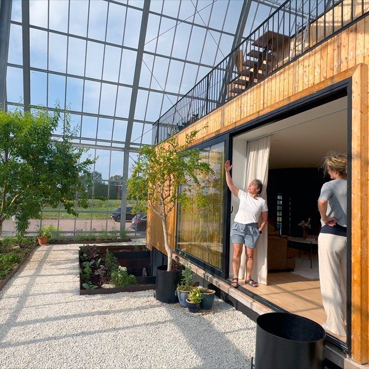 A wooden house with glass doors situated inside a large glass greenhouse structure.
