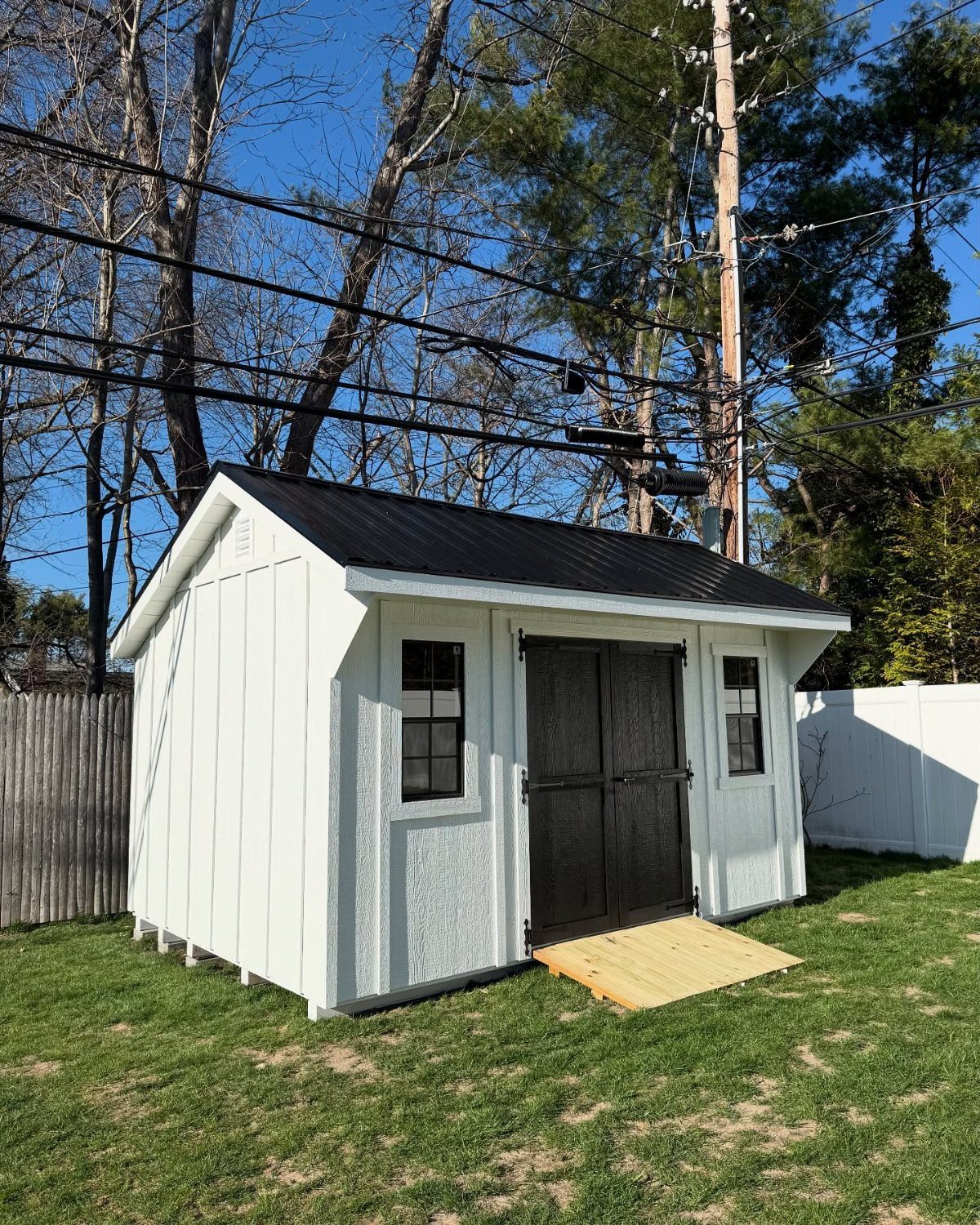 A white modern farmhouse style shed with black doors and trim sitting on a green lawn.